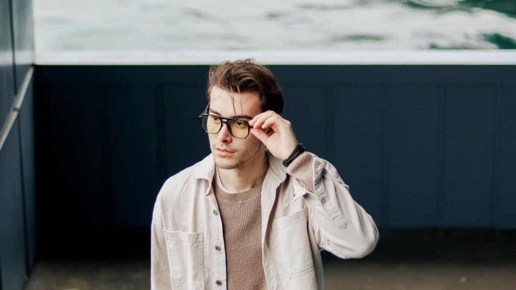 A man adjusting his glasses while standing near a wall.