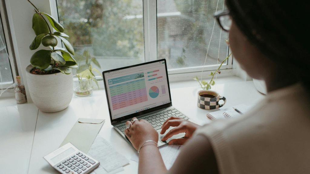 A person wears glasses and works on a laptop showing a spreadsheet by a window.