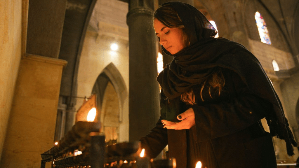 A woman wearing a headscarf lights a candle inside a dimly lit church.