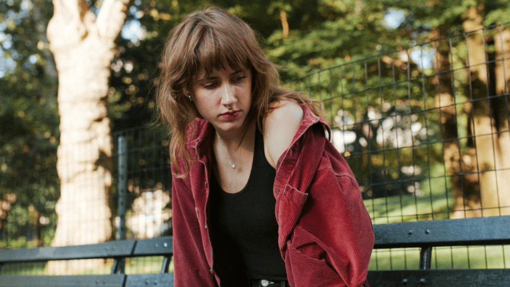 A thoughtful young woman with auburn hair sits alone on a park bench outdoors.