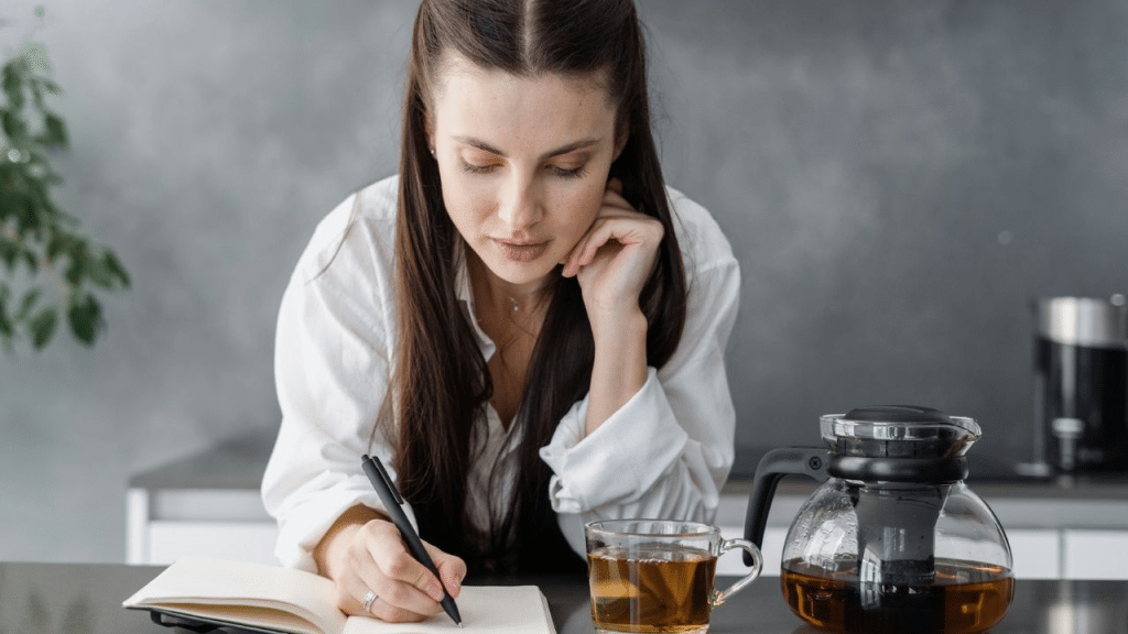 A thoughtful young woman writes in a notebook with a teacup and pot nearby.