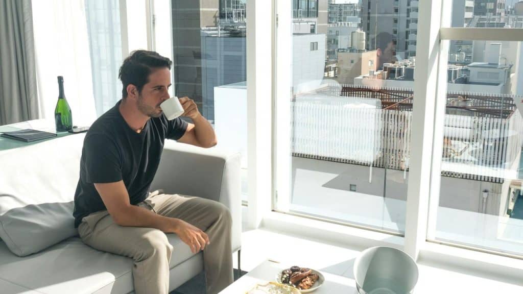 A man sitting on a couch drinking coffee by a large window.