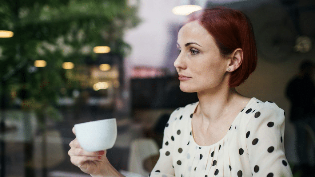 A woman with red hair holds a coffee cup and looks out a window thoughtfully.