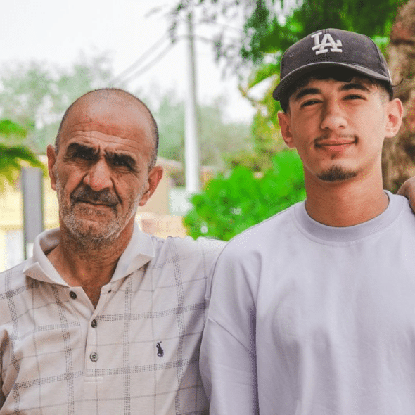 Older man and younger man, with the younger man wearing a hat, posing outdoors.