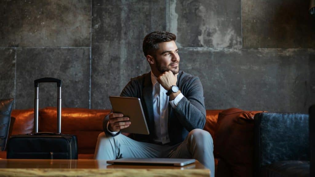 A man sitting on a couch holding a tablet with a suitcase beside him.