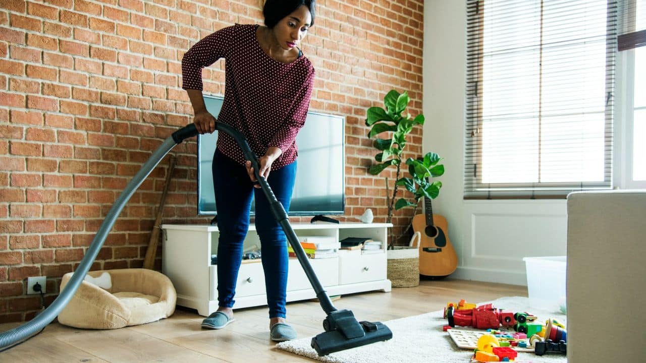 A woman in jeans is vacuuming a rug in a living room with toys on the floor.