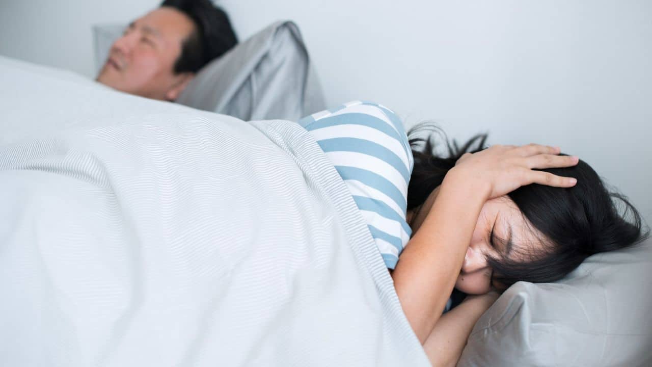 A couple lies in bed, one person is awake with their hand on their head and the other is asleep.