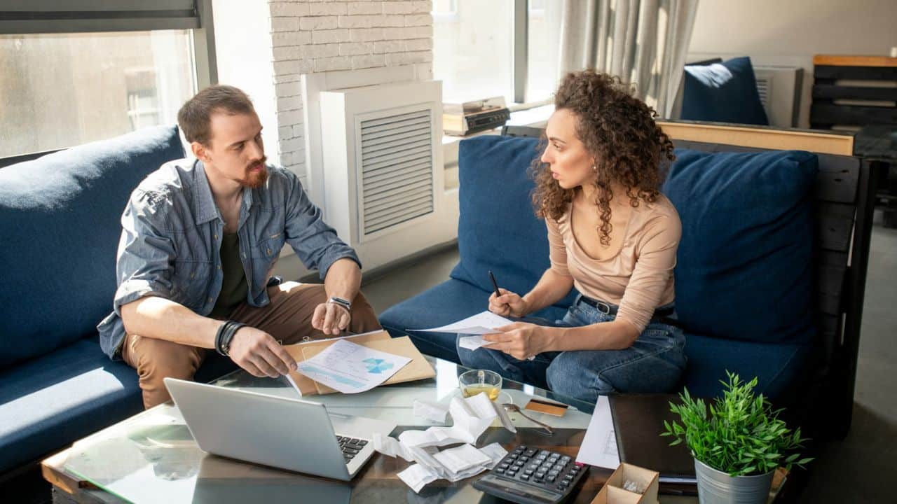 A man and woman sit on a sofa, looking at papers on a table with a laptop.