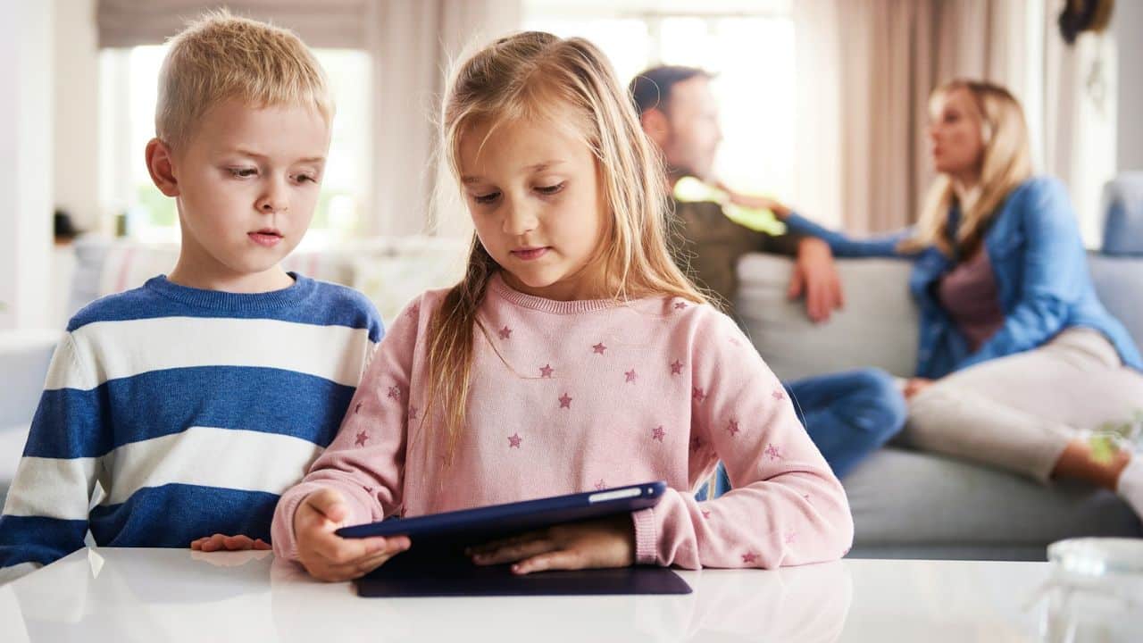 Two children look at a tablet while their parents sit on a sofa in the background.