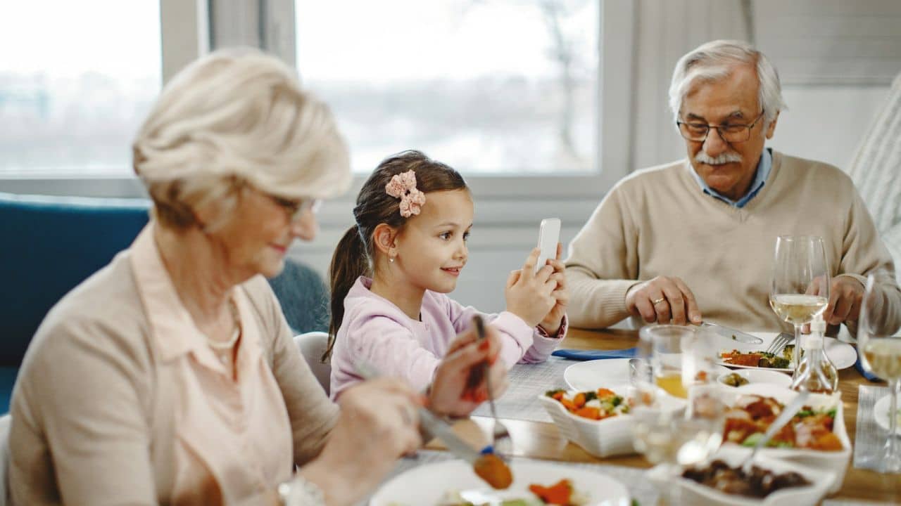 A young girl at a dinner table takes a picture of her smiling grandmother with a phone.