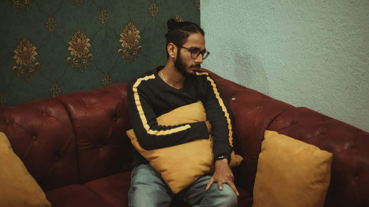 A man with glasses and a man bun sits on a red sofa, hugging a yellow pillow.