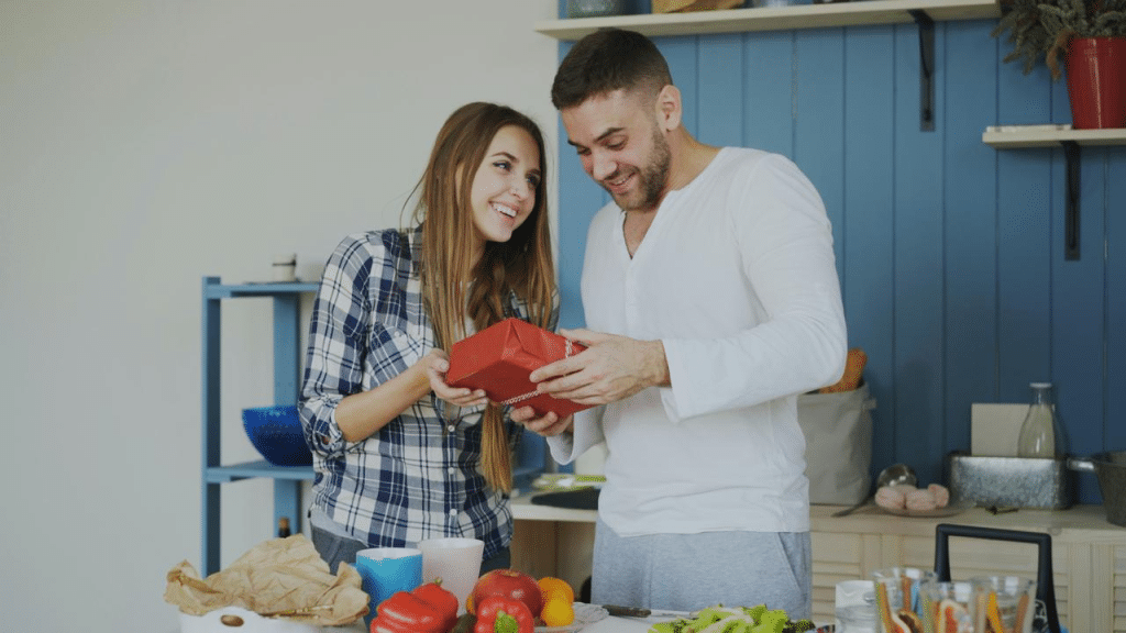 A smiling couple in a kitchen holding a wrapped gift together.