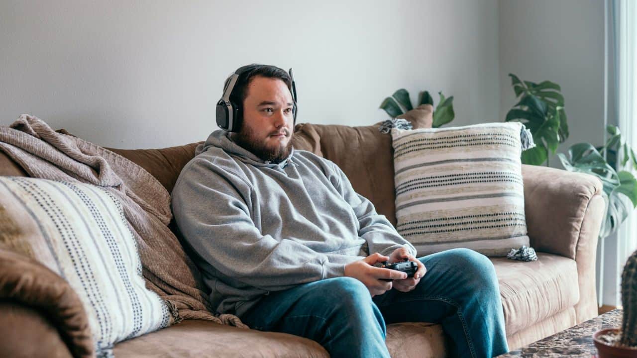 A man with headphones on sits on a sofa, holding a video game controller.