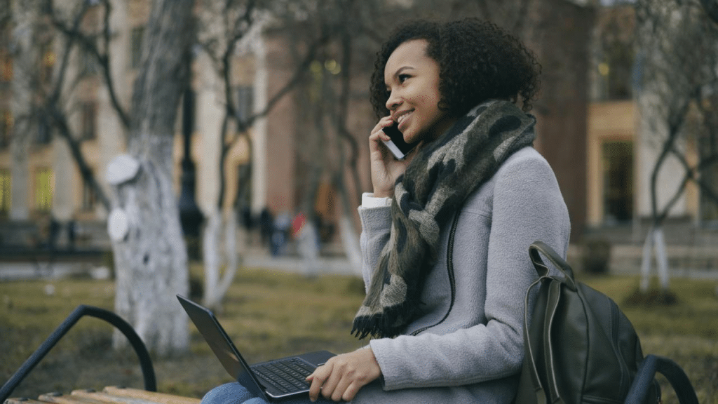 A woman sitting on a bench outdoors talking on the phone with a laptop on her lap.