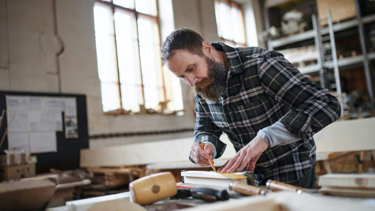 A man with a beard in a plaid shirt carefully measures wood with a pencil in a workshop.