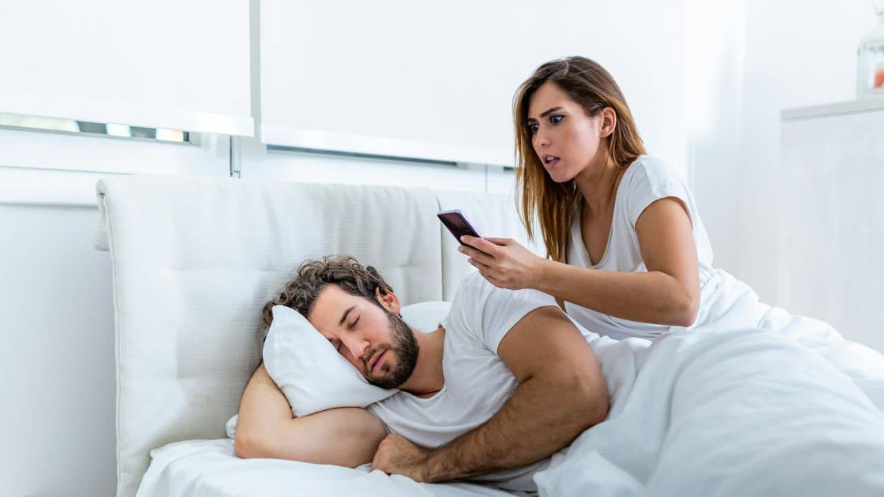 A woman looks at a man's phone while he is asleep in bed.
