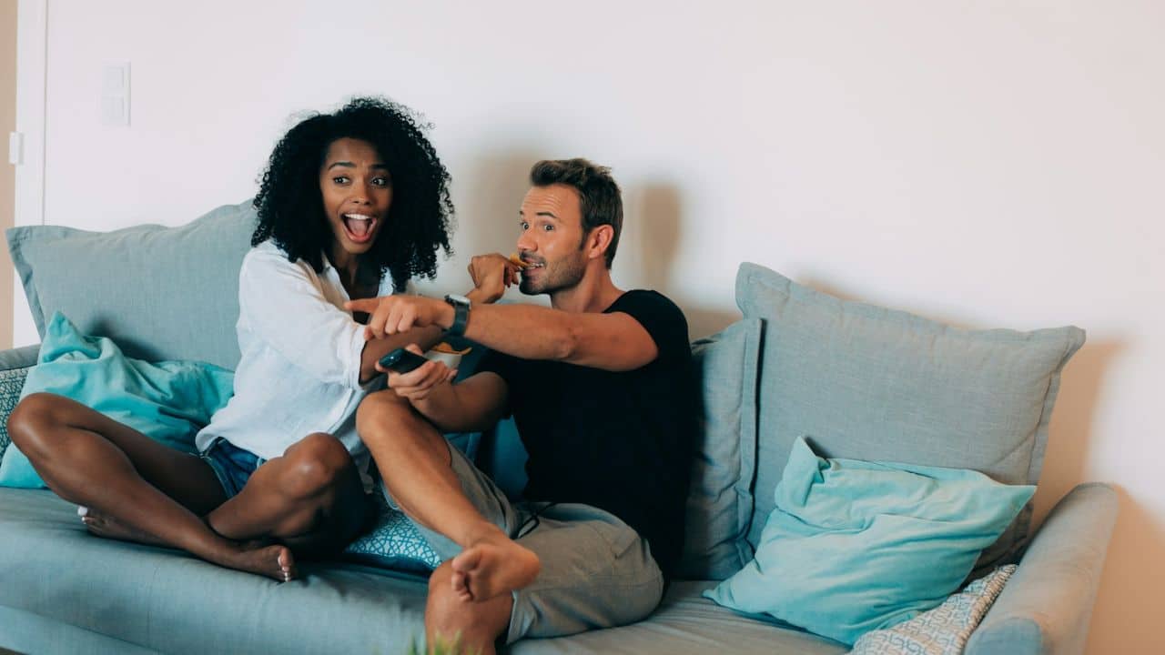 A surprised man and a woman sit on a sofa, looking in different directions.