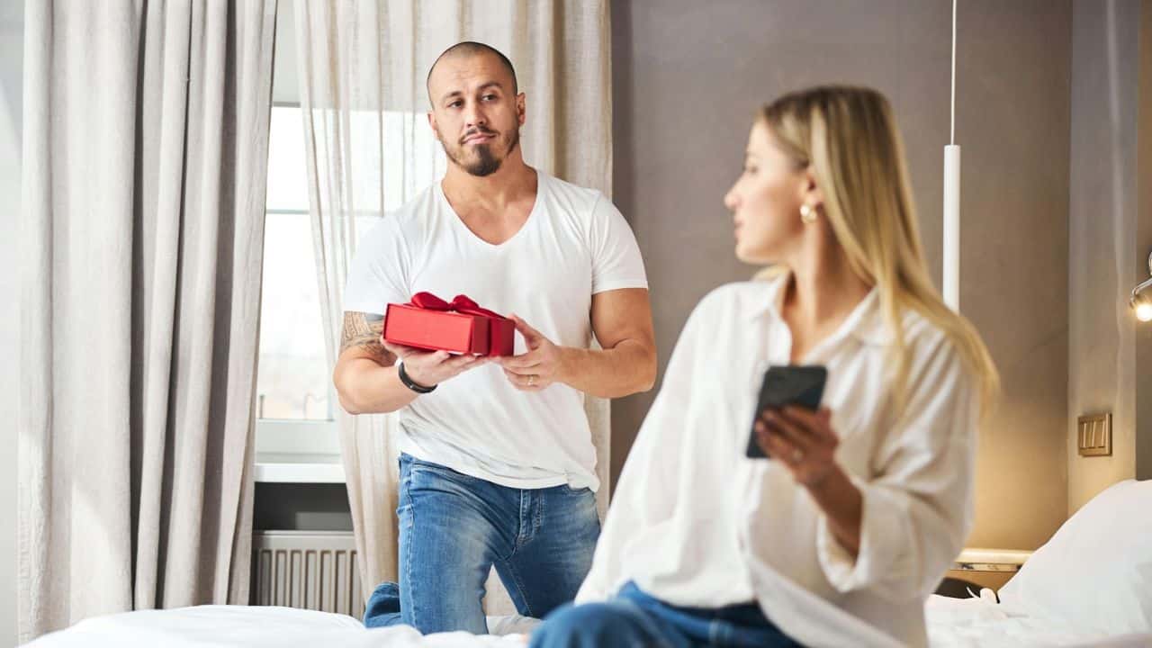 A man on one knee offers a gift box to a woman on a bed who is looking at her phone.
