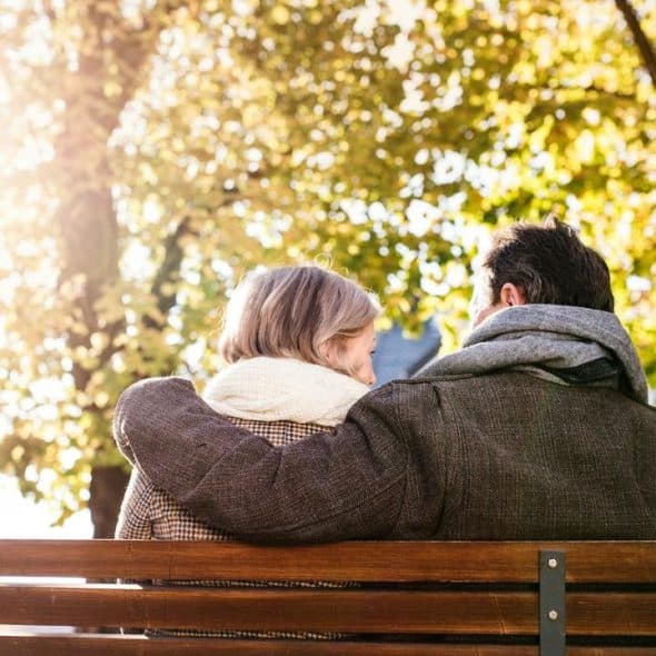 A couple sitting on a park bench with autumn trees in the background.