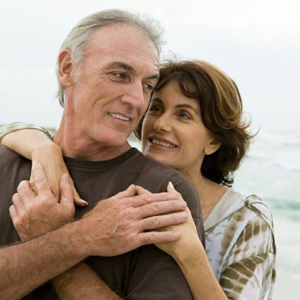 A happy older couple embraces on a beach, looking at each other.