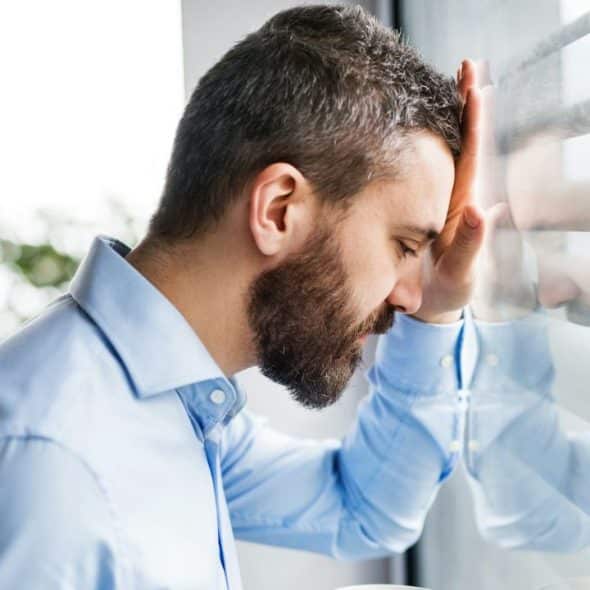A stressed man in a blue shirt leaning his forehead against a window with his eyes closed.