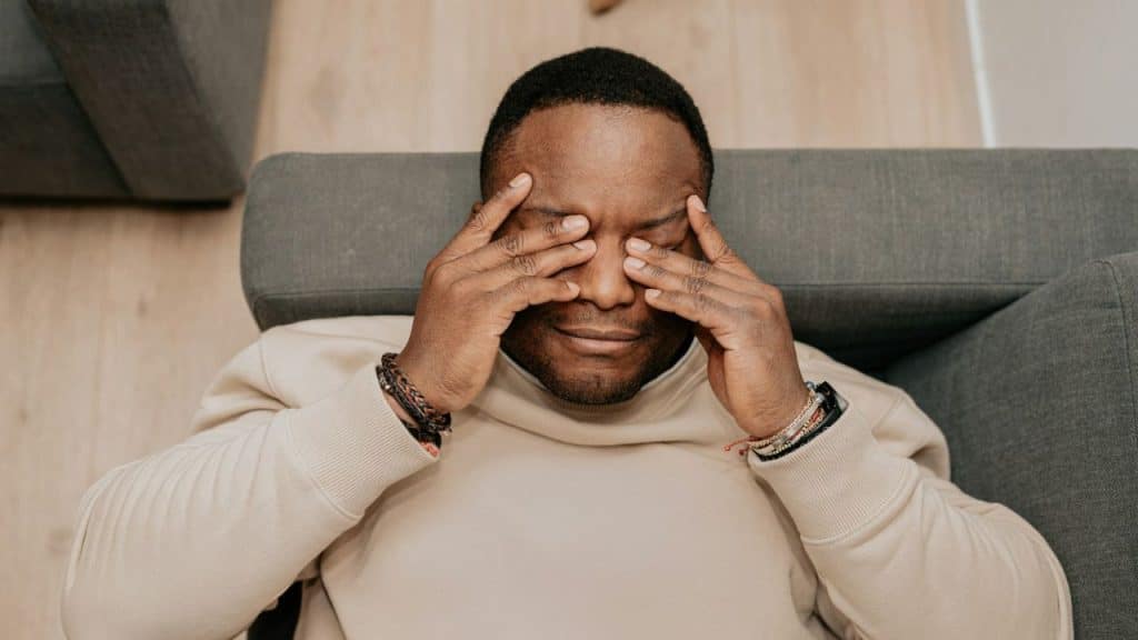 A man lies on a gray couch, rubbing his eyes with both hands.