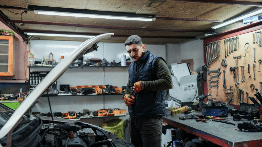 A male mechanic with rubber gloves works on a car in a messy garage.