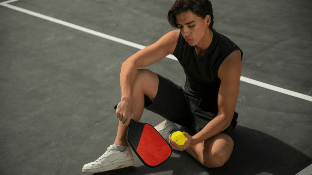 A disappointed man sits on a pickleball court, holding a paddle and a ball.