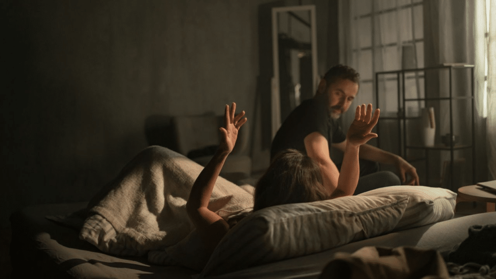 A frustrated woman in bed gestures with her hands as a man looks on, tired and sad.