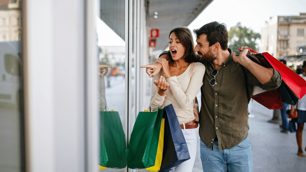 A shocked woman points into a shop window as a man carries shopping bags.