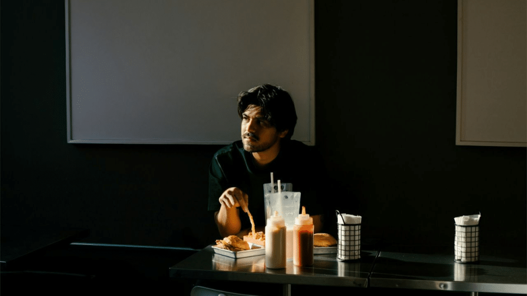A pensive man with dark hair sits alone at a table with food, looking into the distance.