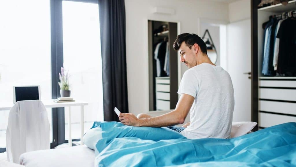 A man sitting on a bed with blue sheets while looking at his phone.