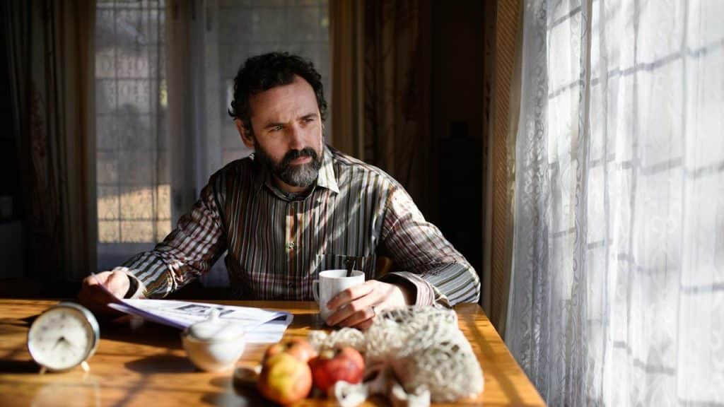 A man sitting at a table with a cup of coffee, looking thoughtfully out the window.