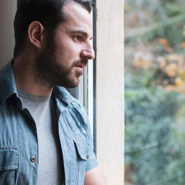 A man in a denim shirt looking out a window thoughtfully..