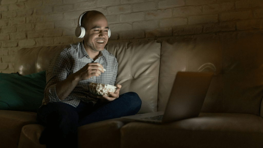 A joyful, balding man in a plaid shirt and headphones eats popcorn while watching something on his laptop.