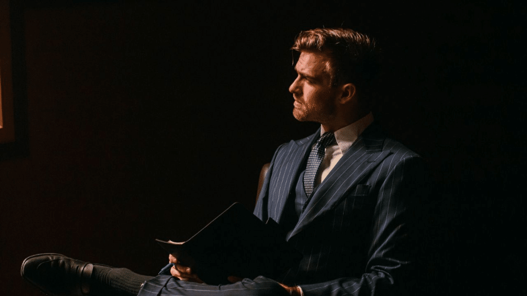 A thoughtful, well-dressed man in a suit sits in a dark room, holding a folder and looking to the side.