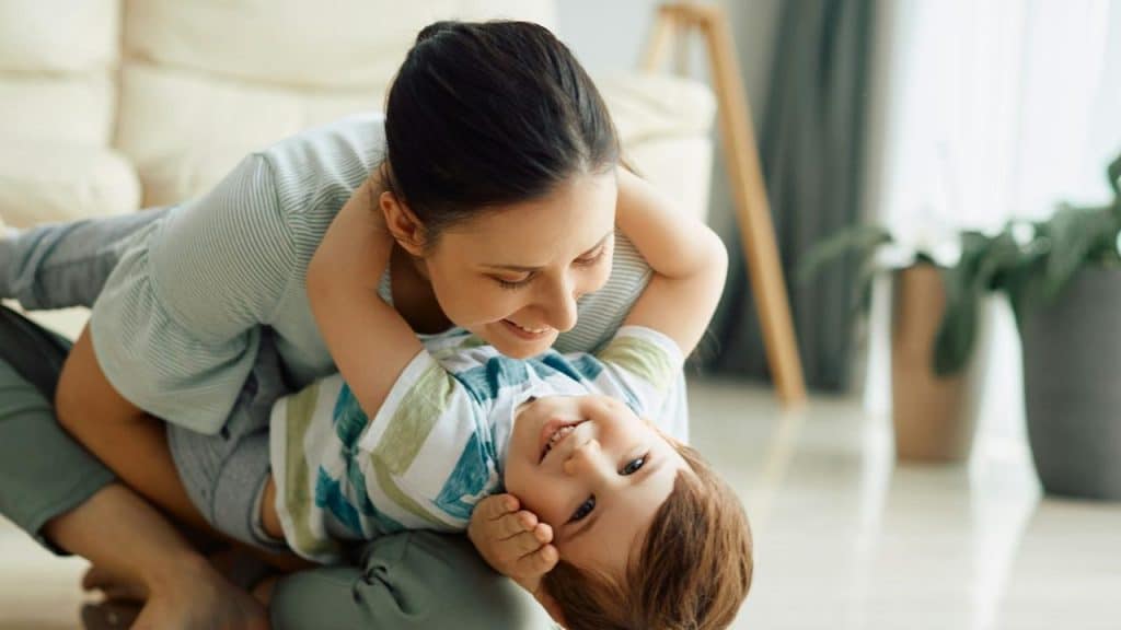 A mother hugs and plays with her smiling child on the floor at home.