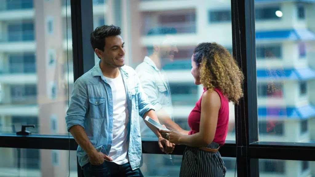 A happy man in a denim shirt smiles at a woman with curly hair, and their reflections can be seen on the window behind them.