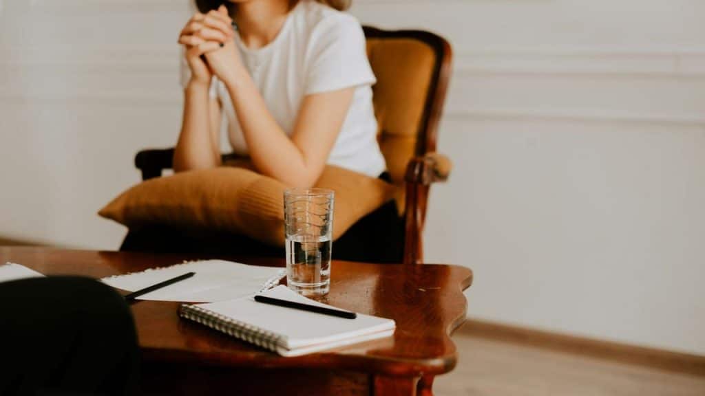 A woman sits with folded hands at a table with notebooks and a glass of water.
