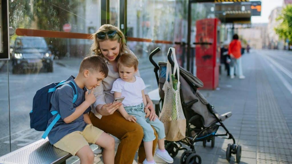A mother sits at a bus stop with her two children, showing them something on her phone.