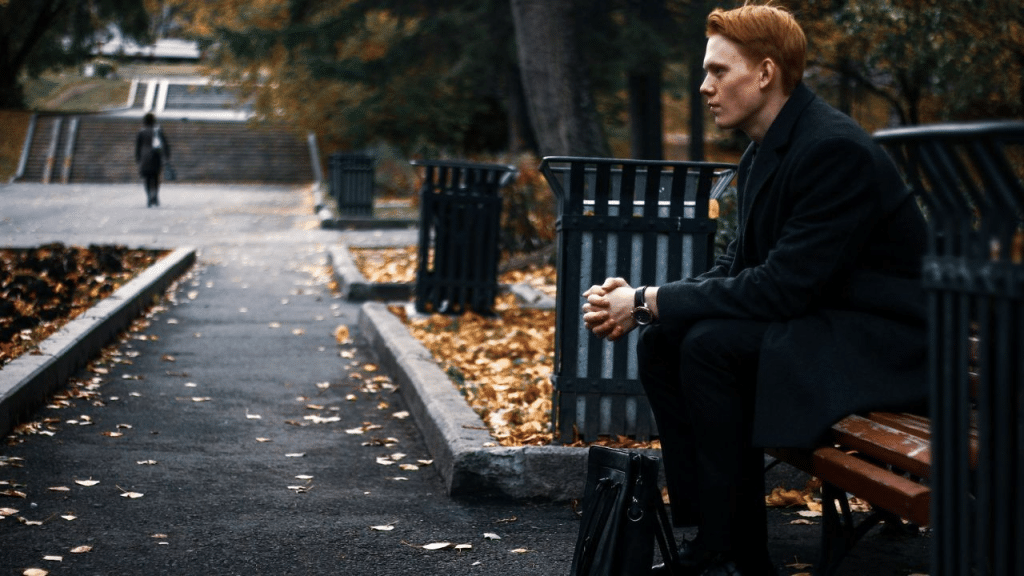A serious-looking man with red hair sits on a park bench, gazing into the distance.