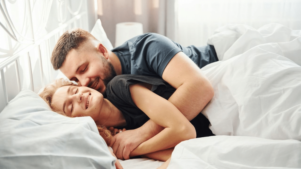 A couple smiles as they hug each other in bed.