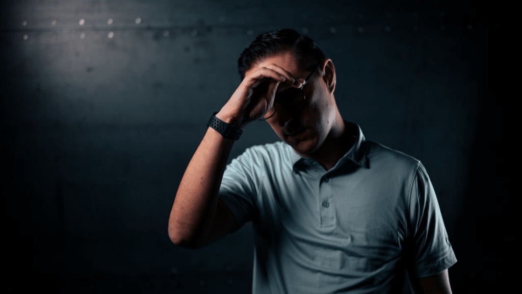 A stressed man in glasses and a light blue polo shirt holds his forehead.