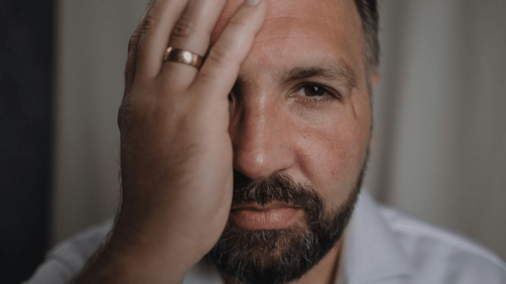 A sad man with a beard and a wedding ring covers his face with his hand.