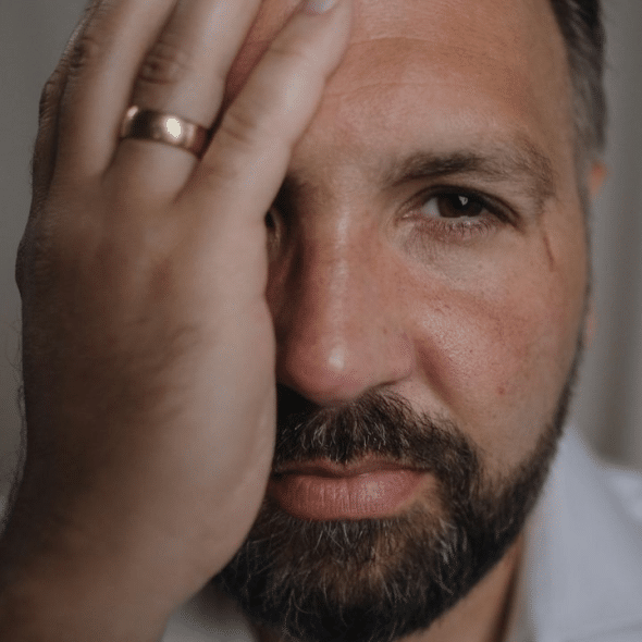 A sad man with a beard and a wedding ring covers his face with his hand.