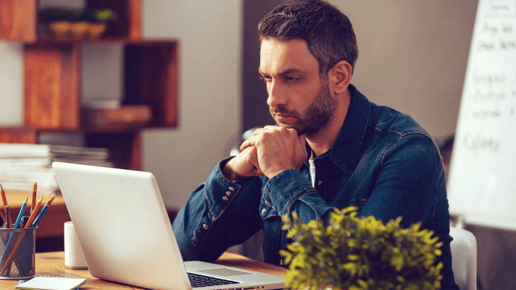 A pensive man with a beard sits at a desk and looks at a laptop.