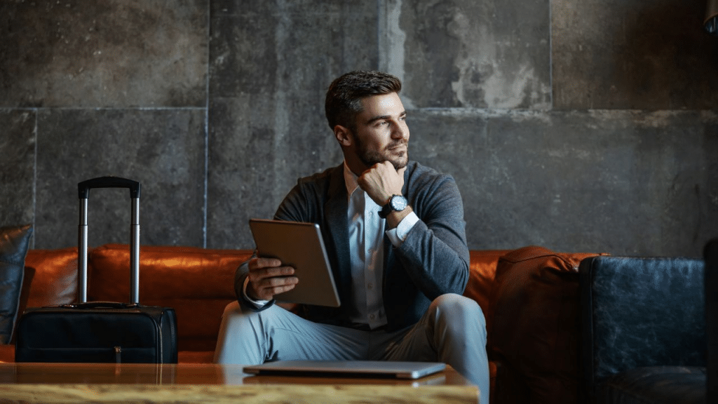 A stylish man sits on a brown leather couch in a modern lobby, holding a tablet and looking to the side.