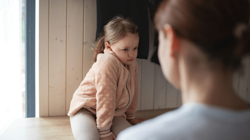 A sad young girl in a fuzzy pink jacket looks away from a woman.