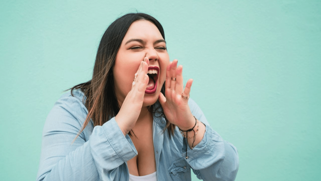 A person with dark hair yells and holds their hands to their mouth.