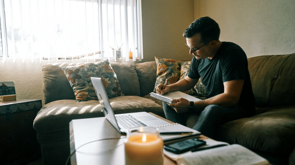 A focused man in glasses and a dark t-shirt writes in a notebook.